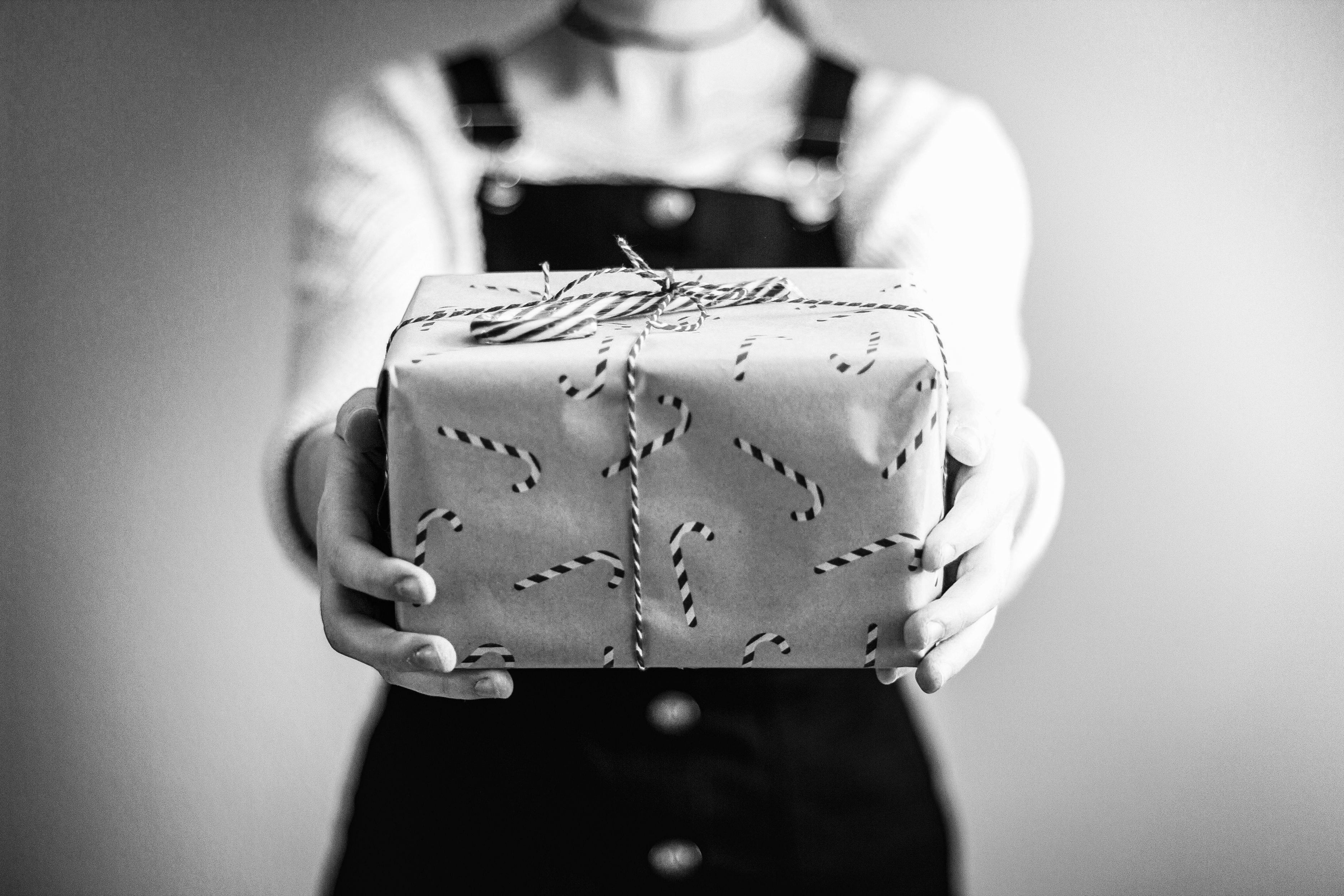 Black and white photo of a person holding out a gift wrapped in candy cane patterned paper, tied with a string.