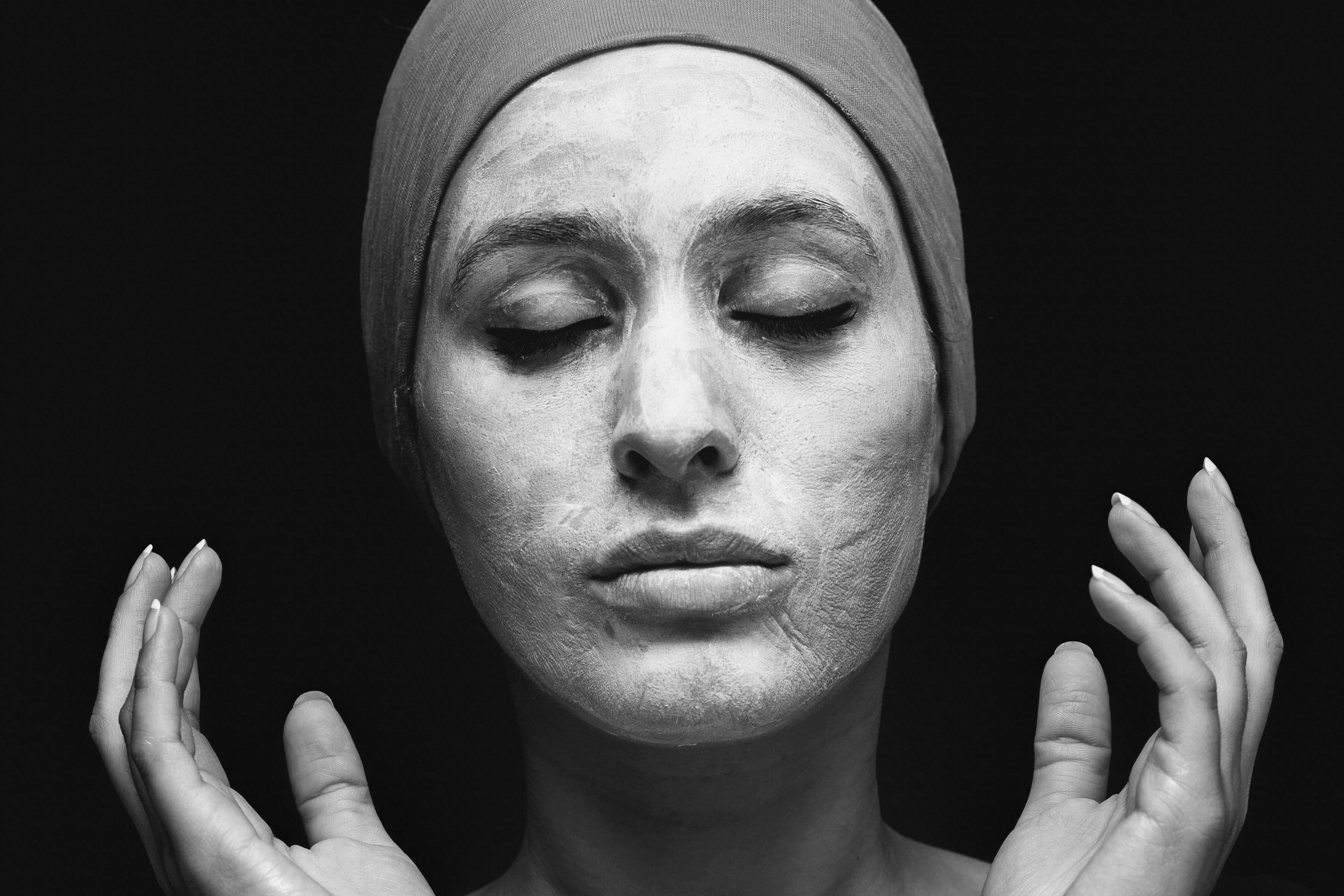 Serene woman with a clay mask and head wrap, eyes closed, against a black background.