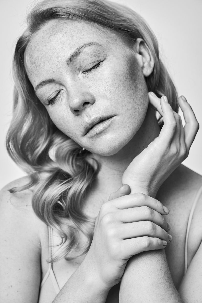 Black and white portrait of a serene woman with closed eyes, gently touching her face and neck with her hands.
