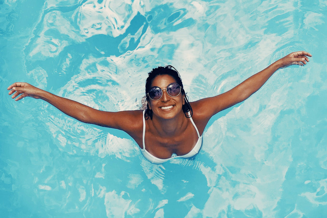 Smiling woman in sunglasses enjoying a swim in a bright blue pool, arms outstretched.