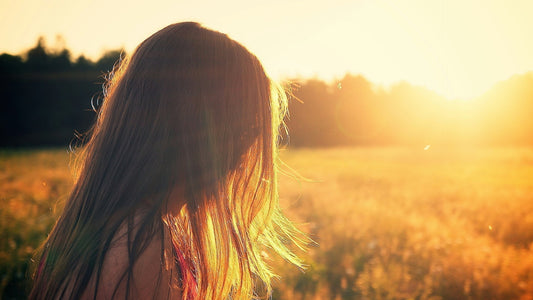A woman with long hair standing in a sunlit field, facing the golden sunset, symbolizing the warmth and beauty of spring sunshine.