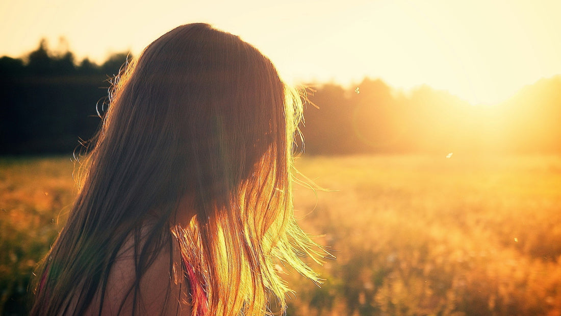 A woman with long hair standing in a sunlit field, facing the golden sunset, symbolizing the warmth and beauty of spring sunshine.